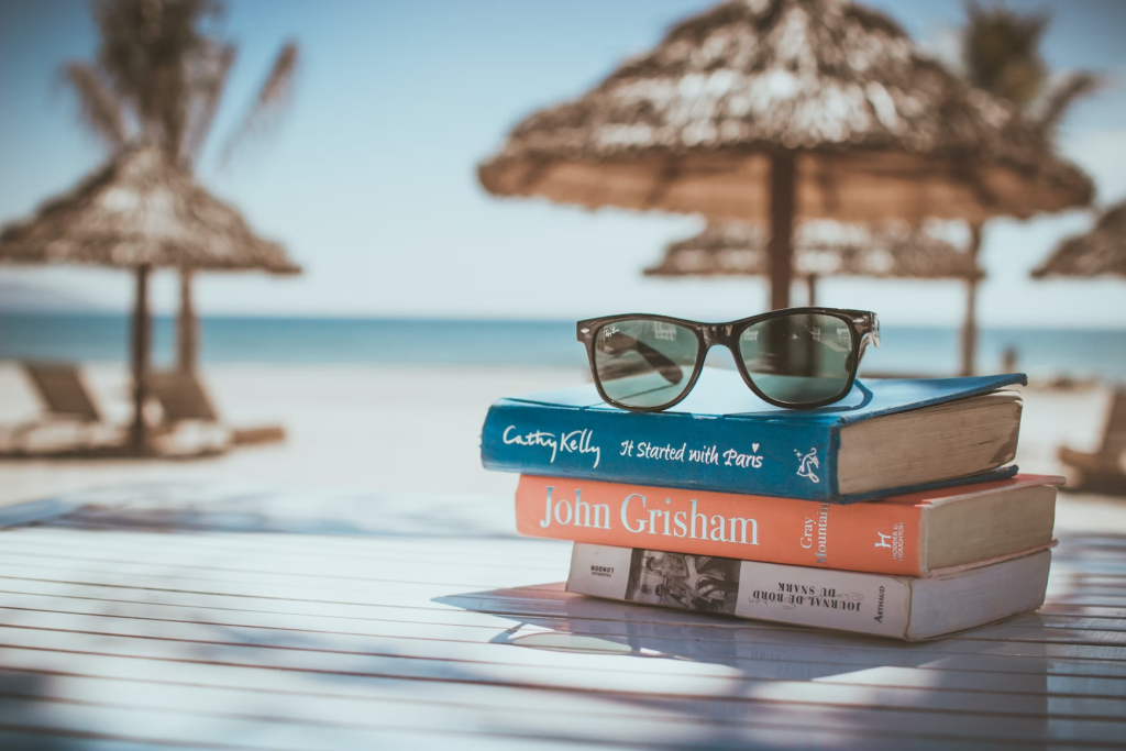 A pile of books with a pair of glasses on  their top, placed on a table which is on a beautiful beach. Start with some easy knowledge to DIY a travel plan!