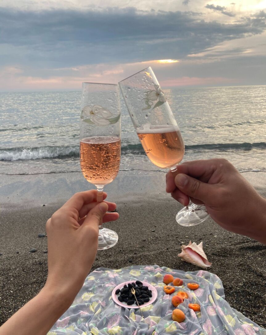 A wine glass rests on the sandy beach, illuminated by the radiant colors of a stunning sunset, creating a serene and picturesque coastal scene