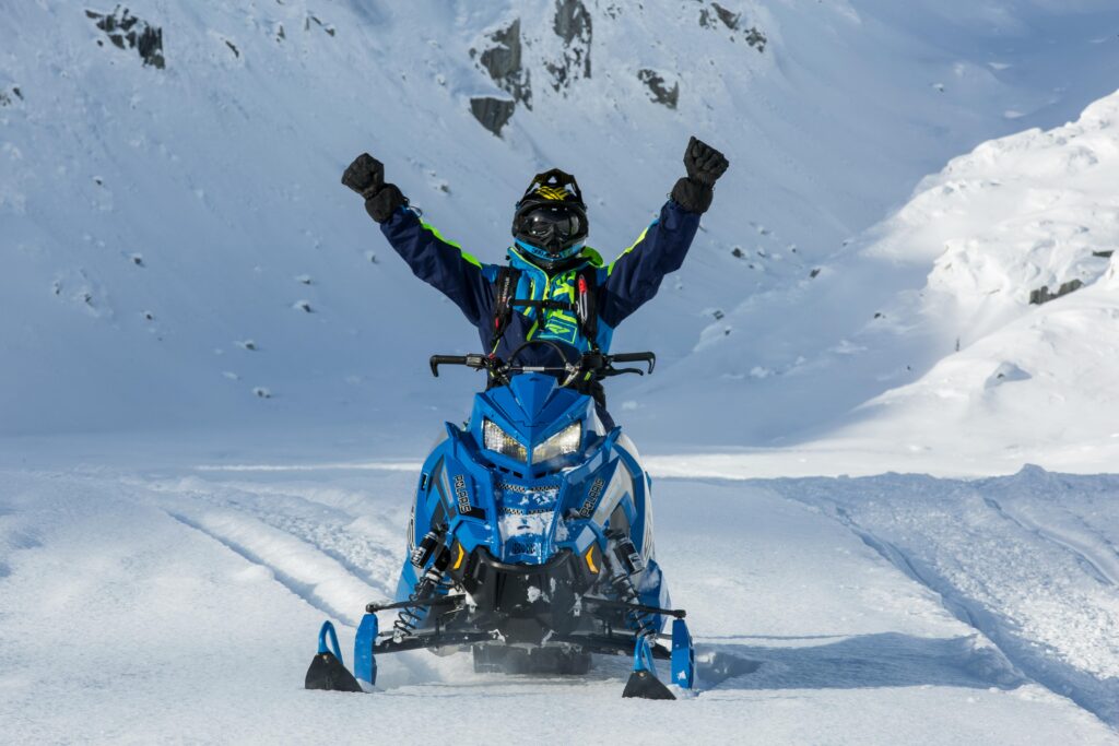 Man raises both hands while riding a snowmobile through a snowy mountain landscape, expressing excitement and adventure. Beginners guide to layering for extreme winter weather conditions